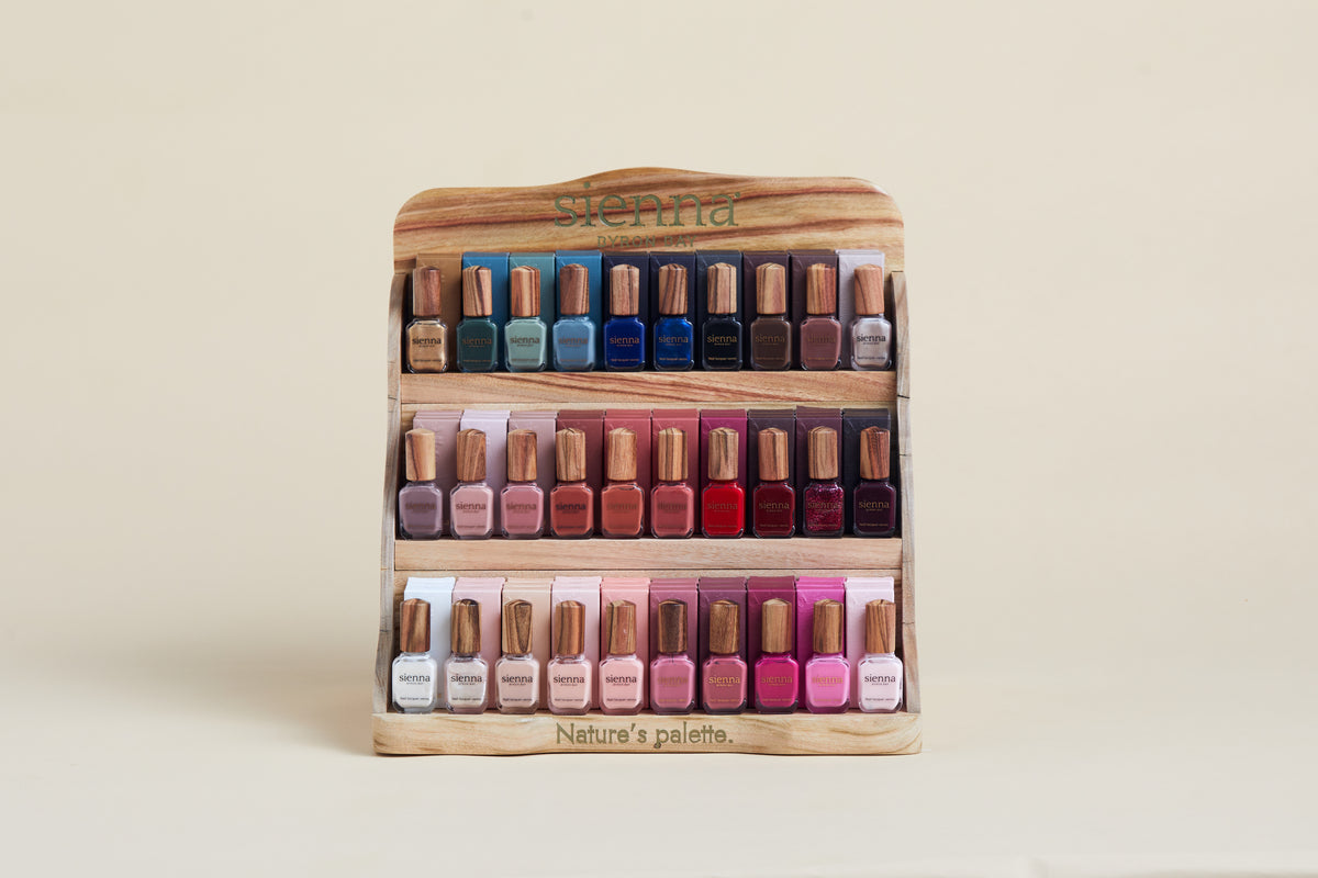 A wooden three-tier nail polish display stand filled with assorted nail polishes in various colors, set against a plain beige background. The brand name 'sienna' is visible on the top of the stand, and 'Nature's palette' is printed on the bottom.
